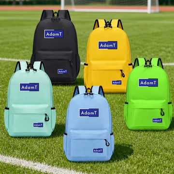 Four backpacks in black, yellow, green, and light blue are arranged on a grassy sports field with a soccer goal in the background. Each backpack has a blue rectangular patch on the front with the brand name 'AdamT' printed on it.