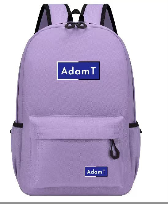 Four backpacks in black, yellow, green, and light blue are arranged on a grassy sports field with a soccer goal in the background. Each backpack has a blue rectangular patch on the front with the brand name 'AdamT' printed on it.